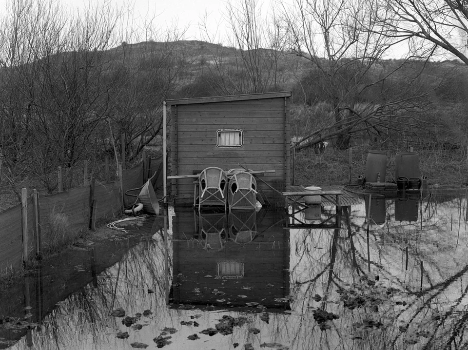 A small timber shed and garden chairs reflected in water surrounding the garden.